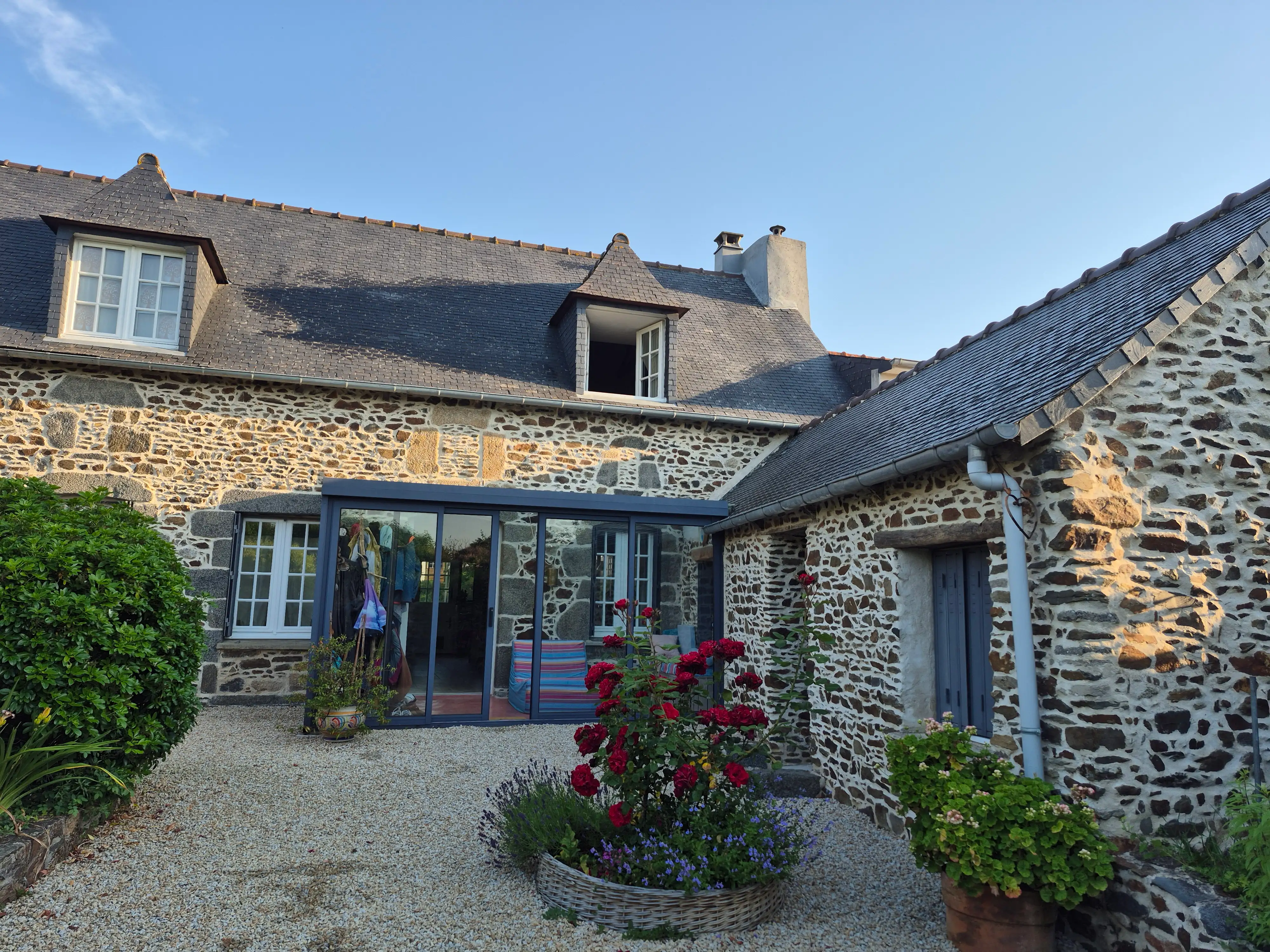 une vue extérieure de la maison en pierre typique de Bretagne, avec la chambre d'hôte à droite, la petite allée de de galets et son décor fleuri